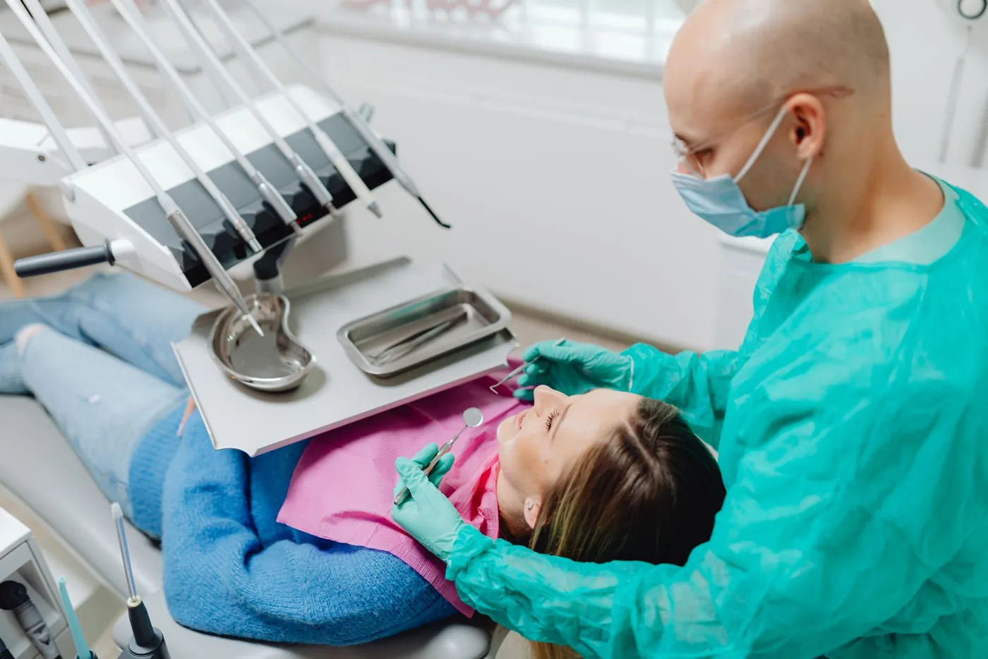 A dentist in scrubs examining a seated patient's teeth with a dental mirror in a bright, modern dental clinic